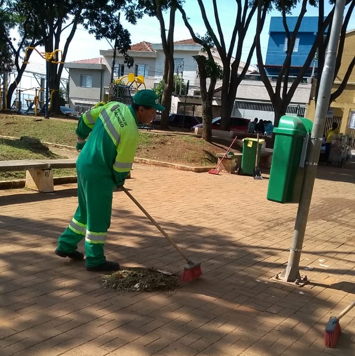 Funcionário da Subprefeitura Ipiranga de costas, vestindo um uniforme verde e boné verde escuro, realizando o serviço de varrição em uma praça